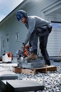 A construction worker using a power cutter on concrete blocks outside a modern house.