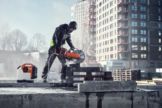 Construction worker using a power cutter on a construction site with buildings in the background.
