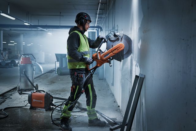 A construction worker using a power cutter on a concrete wall in an indoor construction site.