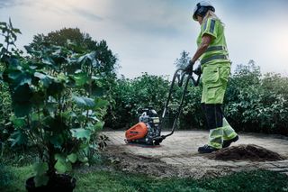 Tányértömörítőt működtető személy egy növényzettel körülvett kerti ösvényen.