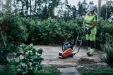 Een persoon die een trilplaat gebruikt op een verhard buitenterrein omgeven door groen.