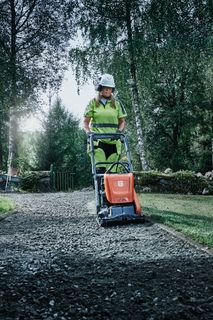 A person operating a compact plate compactor on a gravel path in a garden setting.
