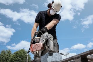A construction worker using a power cutter to cut stone outdoors, wearing protective gear.