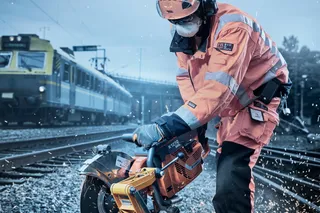 A construction worker using a power cutter on railway tracks, with a train in the background.