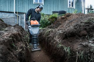 Personne utilisant un compacteur de sol dans une tranchée à proximité d’un bâtiment.