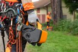 Uma pessoa a segurar um produto Husqvarna com equipamento de escalada, enquanto outra pessoa está em segundo plano num jardim.