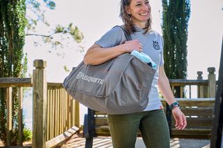 A person carrying a large Husqvarna bag outdoors, near a wooden fence and trees.