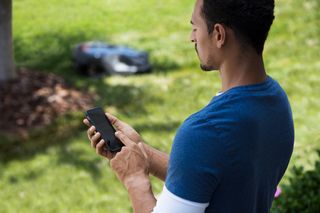 Людина, яка використовує смартфон для керування роботизованою газонокосаркою в саду.