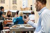 A person sitting at a cafe table holding a smartphone, with a coffee cup and croissant nearby.