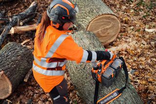 Uma pessoa com equipamento de trabalho, transportando um produto Husqvarna alimentado a bateria, de pé ao lado de cortar troncos de árvores.