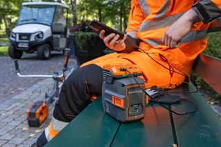 A person sitting on a bench using a smartphone with a Husqvarna battery and trimmer nearby.
