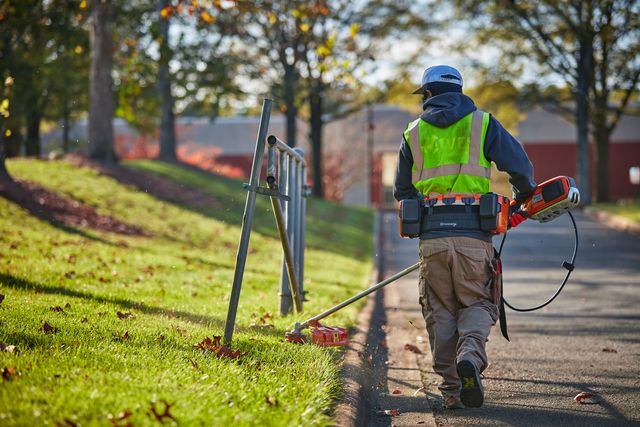 A person using a trimmer to maintain grass along a roadside, wearing work gear.