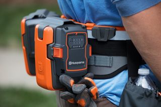 En person, som bærer et bælte med to Husqvarna-batteripakker påmonteret, og som holder arbejdshandsker.