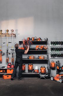 Persona, kas organizē dažādus dārzkopības un celtniecības instrumentus plauktos, ar baterijām, kas uzstādītas pie sienas.