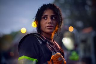 Una persona que lleva ropa de trabajo con rayas reflectantes y guantes, de pie al aire libre al anochecer.