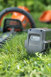 Una batteria Husqvarna posizionata sull'erba con un trimmer sullo sfondo.