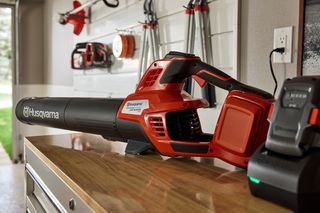 A Husqvarna leaf blower and battery charger on a wooden table in a garage setting.