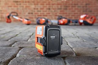 Close-up of a Husqvarna battery on a cobblestone surface with blurred garden tools in the background.