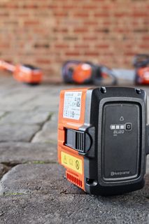 Primo piano di una batteria Husqvarna su una superficie di ciottoli con attrezzi da giardino sfocati sullo sfondo.