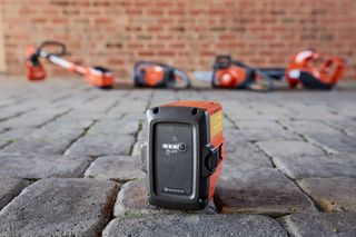 Lähikuva Husqvarnan akusta mukulakivipinnalla ja taustalla erilaisia Husqvarna-työkaluja.