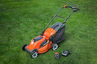 Une tondeuse à batterie Husqvarna sur une pelouse herbeuse avec une batterie et un chargeur à côté.