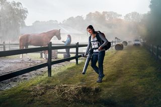 一个人使用吹叶机在马厩区域清理干草,背景是一匹马和另一个人。