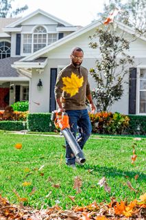 Une personne utilise un souffleur de feuilles pour dégager les feuilles mortes d’un sentier devant une grande maison.