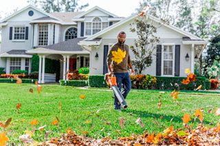 Une personne utilise un souffleur de feuilles pour dégager les feuilles mortes d’un sentier devant une grande maison.