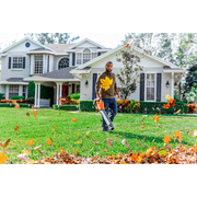 Une personne utilise un souffleur de feuilles pour dégager les feuilles mortes d’un sentier devant une grande maison.