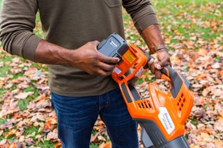 Een persoon die een batterij in een Husqvarna-bladblazer plaatst op een met bladeren bedekt pad.