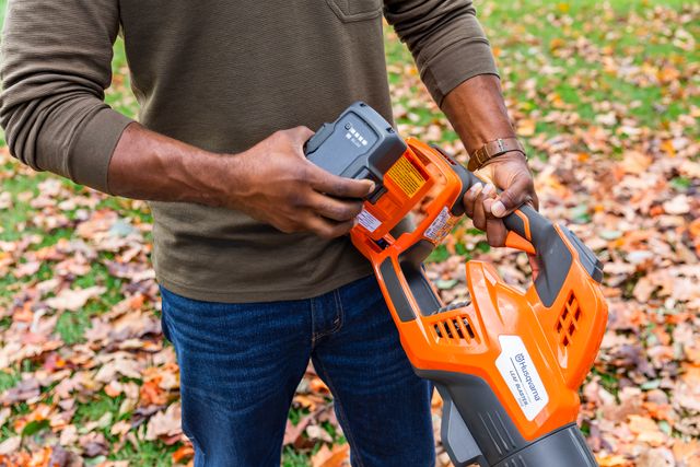 A person inserting a battery into a Husqvarna leaf blower on a leaf-covered path.