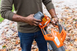 A person inserting a battery into a Husqvarna leaf blower on a leaf-covered path.