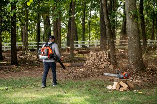 A person using a leaf blower to clear fallen leaves in a wooded area, with a chainsaw resting on a tree stump.