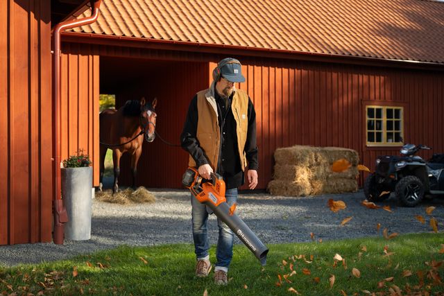 Iemand die een bladblazer gebruikt om gevallen bladeren te verwijderen bij een stal met een paard en een ATV op de achtergrond.