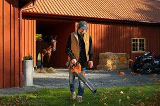 Eine Person, die ein Laubblasgerät benutzt, um heruntergefallene Blätter in der Nähe eines Stalls zu entfernen, mit einem Pferd und einem Quad im Hintergrund.