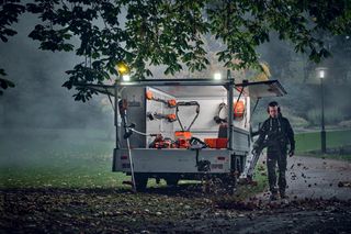 Une personne utilise un souffleur de feuilles pour débroussailler les feuilles mortes dans un parc, avec une remorque contenant divers outils de jardinage.