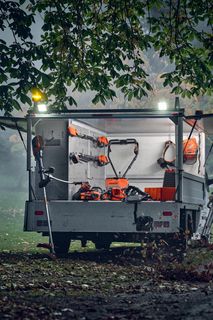 Eine Person, die ein Laubblasgerät benutzt, um heruntergefallene Blätter in einem Park zu beseitigen, mit einem Anhänger, der verschiedene Gartengeräte enthält.