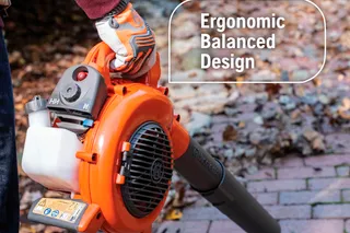 A person holding a leaf blower with an ergonomic balanced design, standing on a brick path with fallen leaves.
