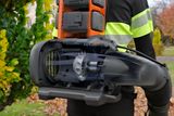 A person holding a leaf blower with visible battery compartments in a park setting.