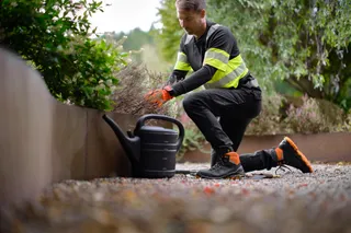 A person in work gear kneeling on a gravel path in a garden setting.