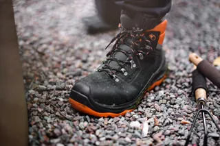 A person wearing work shoes standing on gravel, with gardening tools nearby.