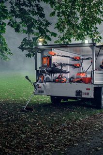 Una persona in piedi accanto a un rimorchio che espone vari attrezzi da giardinaggio in un parco.