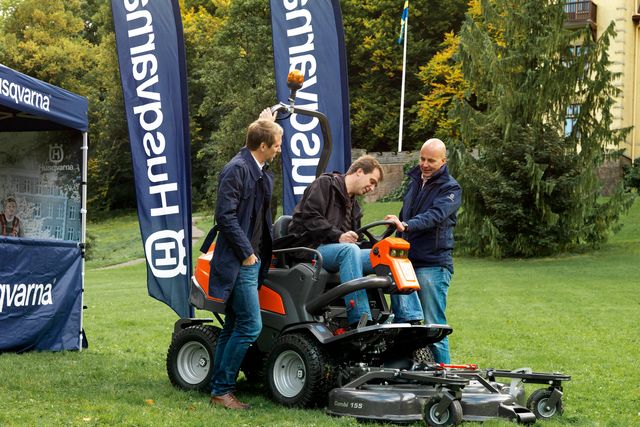 Två personer demonstrerar en Husqvarna åkgräsklippare med klippaggregat i en trädgård.