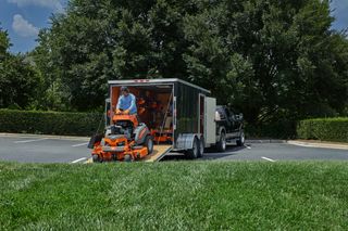 A person unloading a ride-on mower from a trailer in a parking lot.