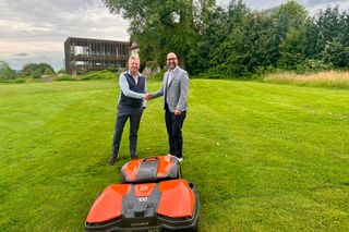 Zwei Menschen schütteln sich auf einer Wiese die Hände mit einem Mähroboter im Vordergrund.