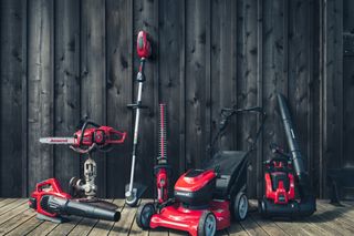 A collection of gardening tools including a chainsaw, hedge trimmer, lawn mower, and leaf blower on a wooden deck.