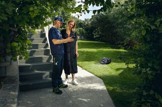 Dos personas de pie en las escaleras de un jardín, observando un robot cortacésped moviéndose sobre el césped.