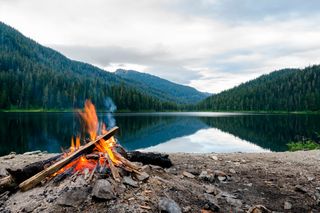 A serene lakeside view with a campfire burning on the shore, surrounded by dense forest.