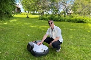 Személy egy kertben, füves területen térdelve egy robotfűnyíróval foglalkozik.