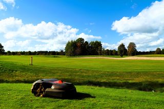 Robotic lawn mower on a large golf course fairway near a sand bunker under a blue sky.